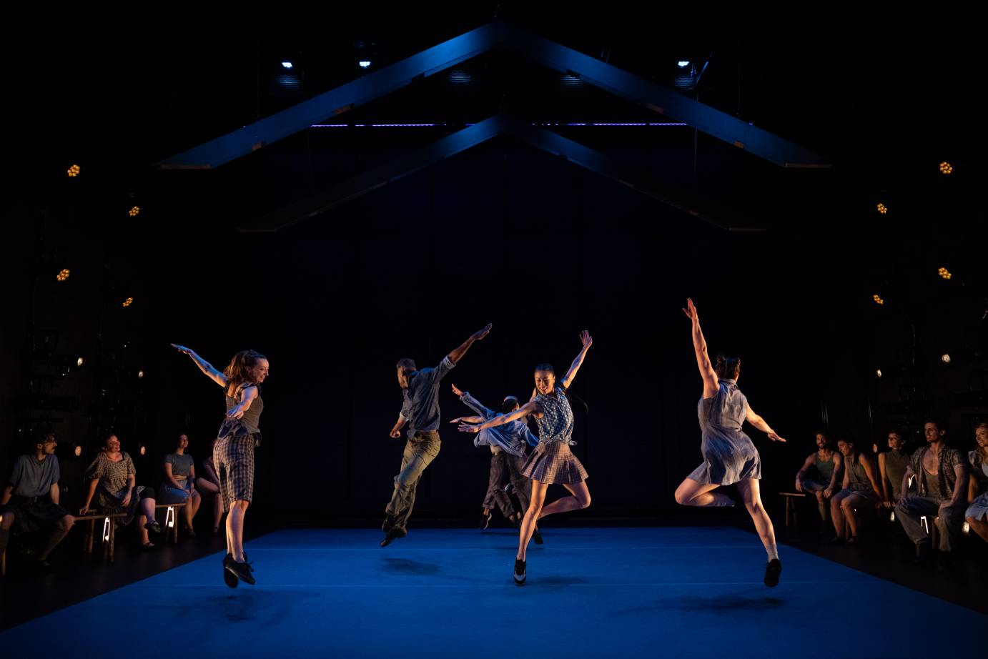 a group of of 5 dancers jump in the air, their arms outstretched. They are having fun while on either side of them groups of friends sitting on benches watch. 